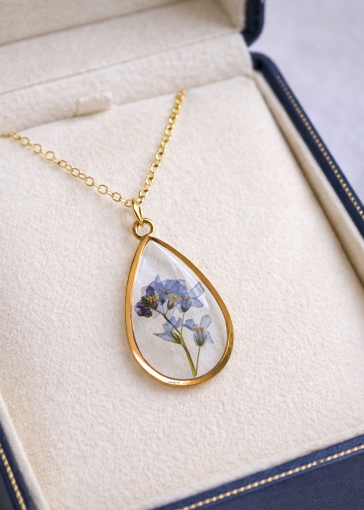 Gold necklace with a teardrop-shaped pendant featuring pressed flowers, displayed on a white box.