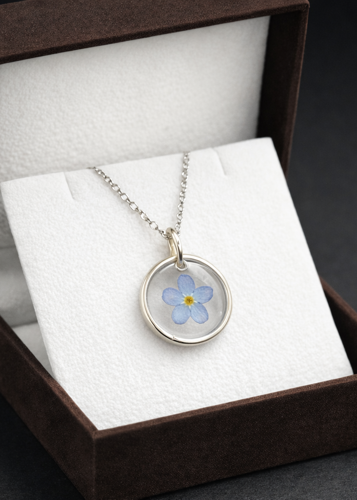 Silver necklace with a clear pendant featuring a blue flower inside a brown box on a dark surface